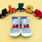 Two gray baby socks with red train designs, arranged side-by-side on a yellow surface with a toy train made of colorful wooden blocks in the background.