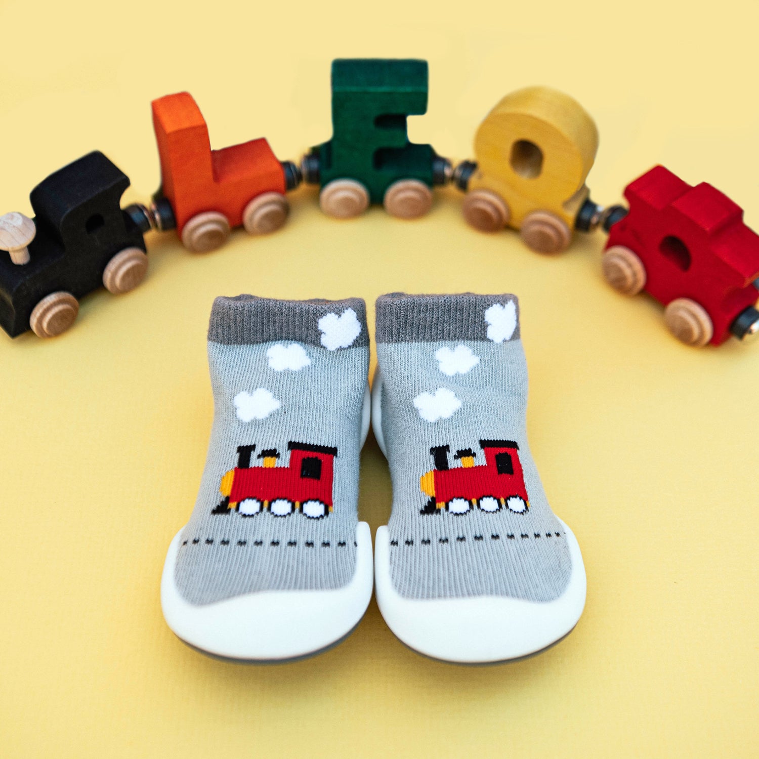 Two gray baby socks with red train designs, arranged side-by-side on a yellow surface with a toy train made of colorful wooden blocks in the background.