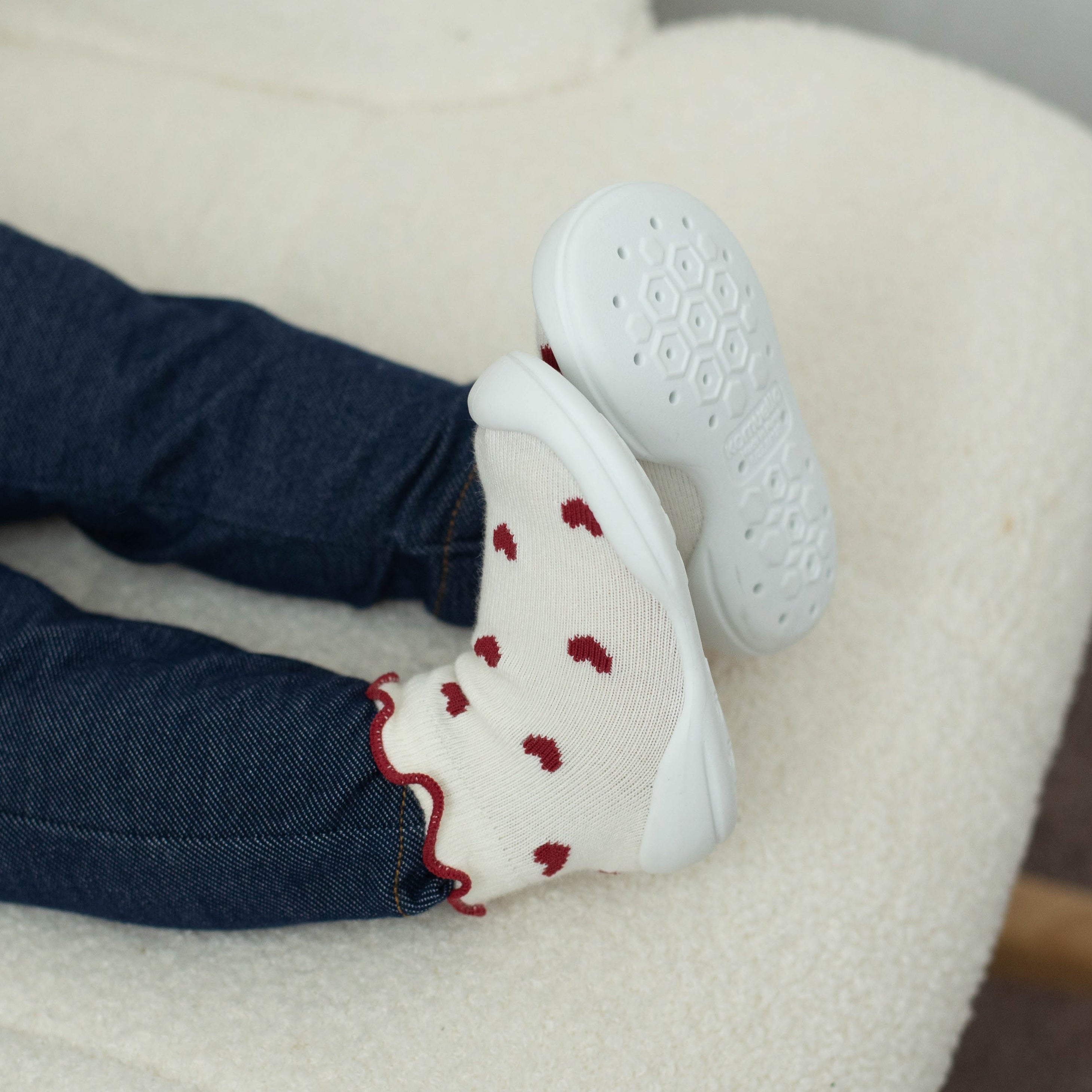 Legs in jeans and white socks with red heart pattern. White shoe sole visible. Fluffy white background.