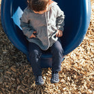 Child sitting on a blue slide, wearing a gray coat, blue pants, and polka dot socks, surrounded by wood chips.
