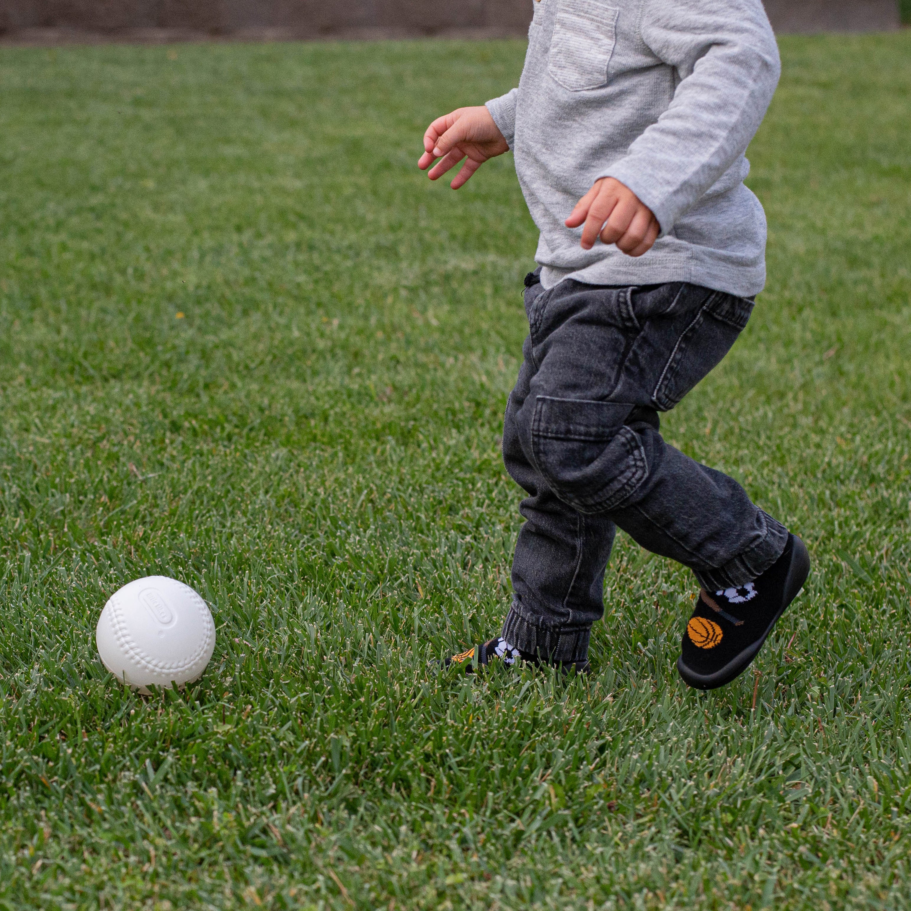 Toddler in gray shirt and dark jeans walking on grass toward a white baseball. The toddler wears black shoes with basketball and other sports designs.