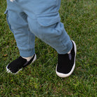 Child wearing blue pants and black and white slip-on shoes standing on green grass.
