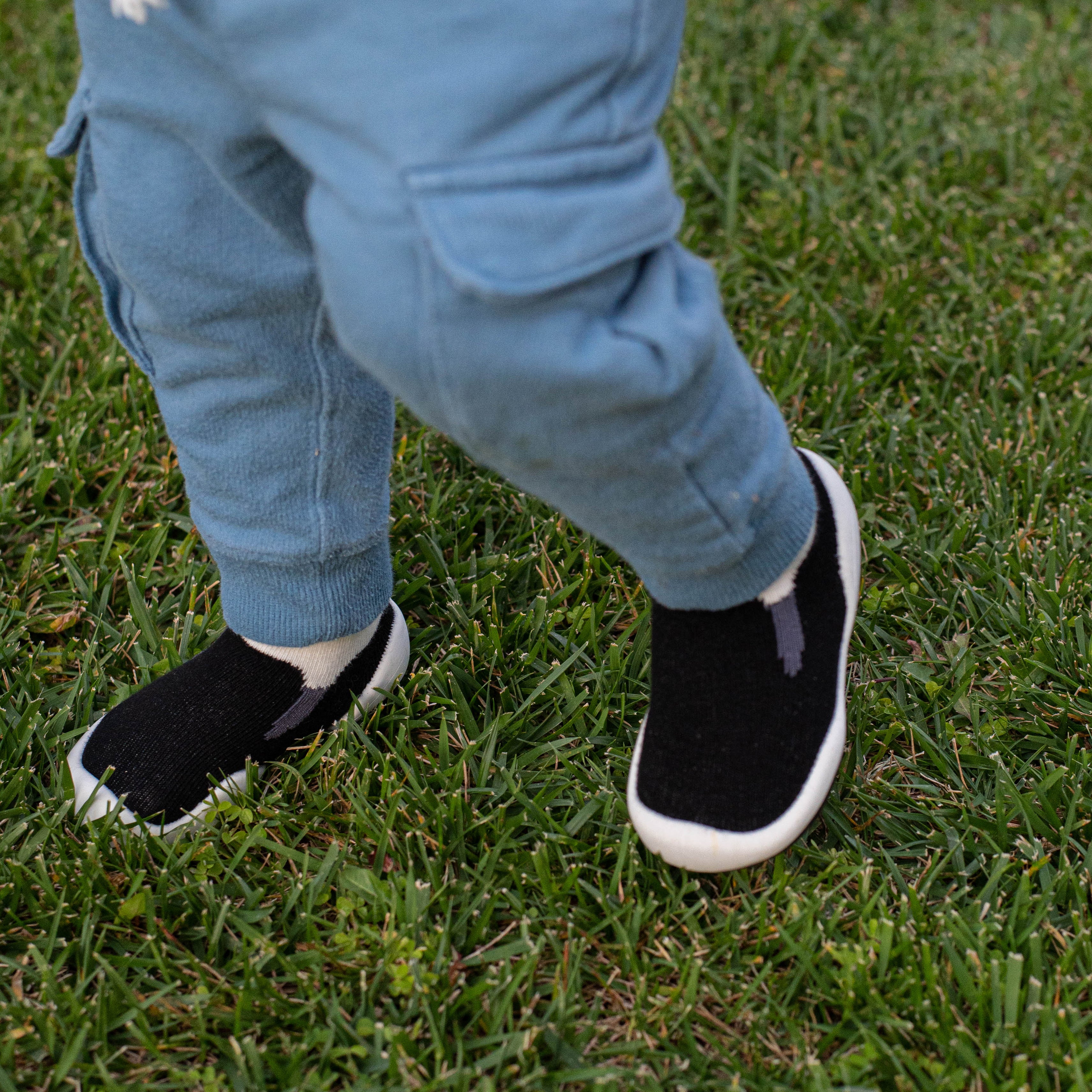 Child wearing blue pants and black and white slip-on shoes standing on green grass.
