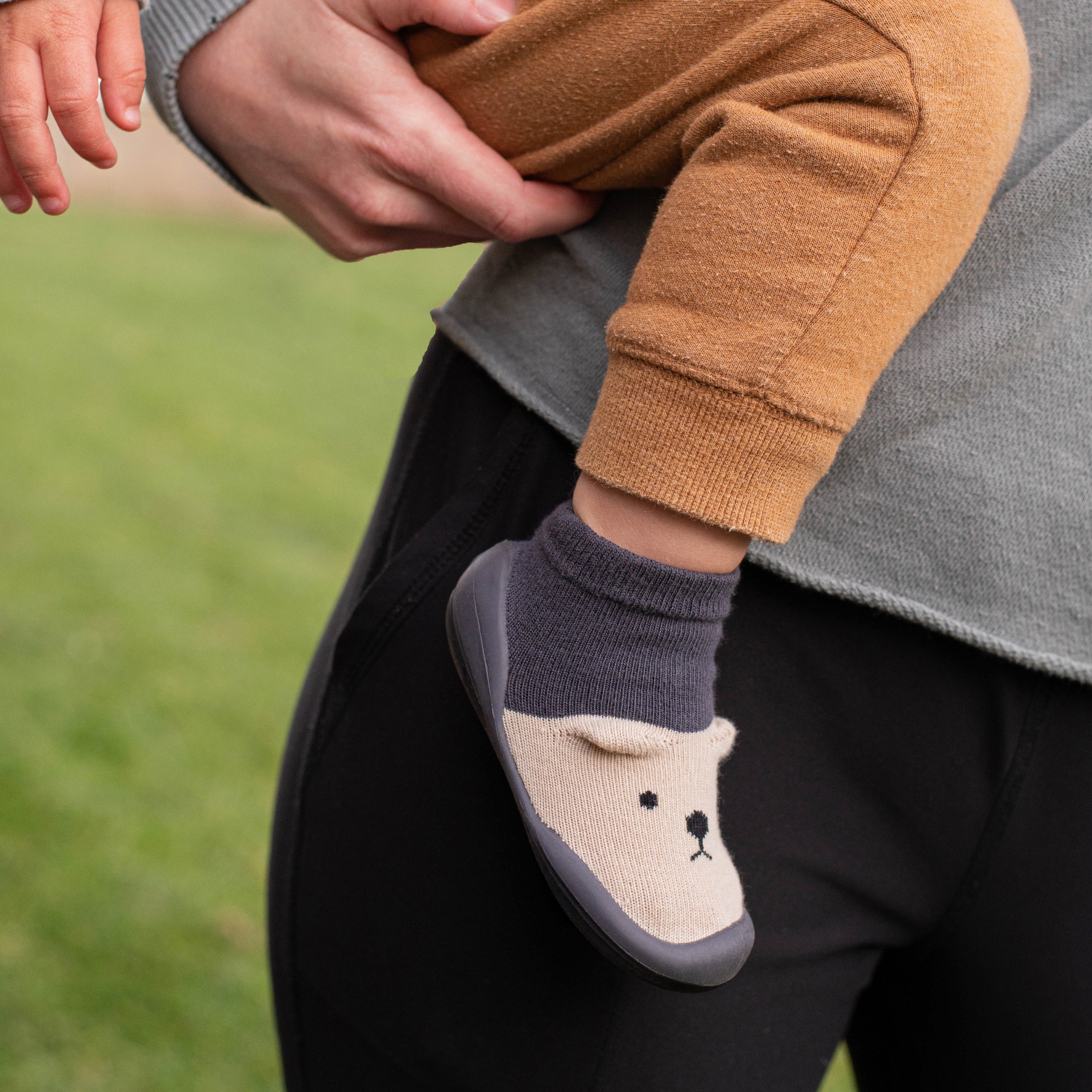 A person is holding a baby wearing brown pants and a gray and beige sock with a bear face on it.