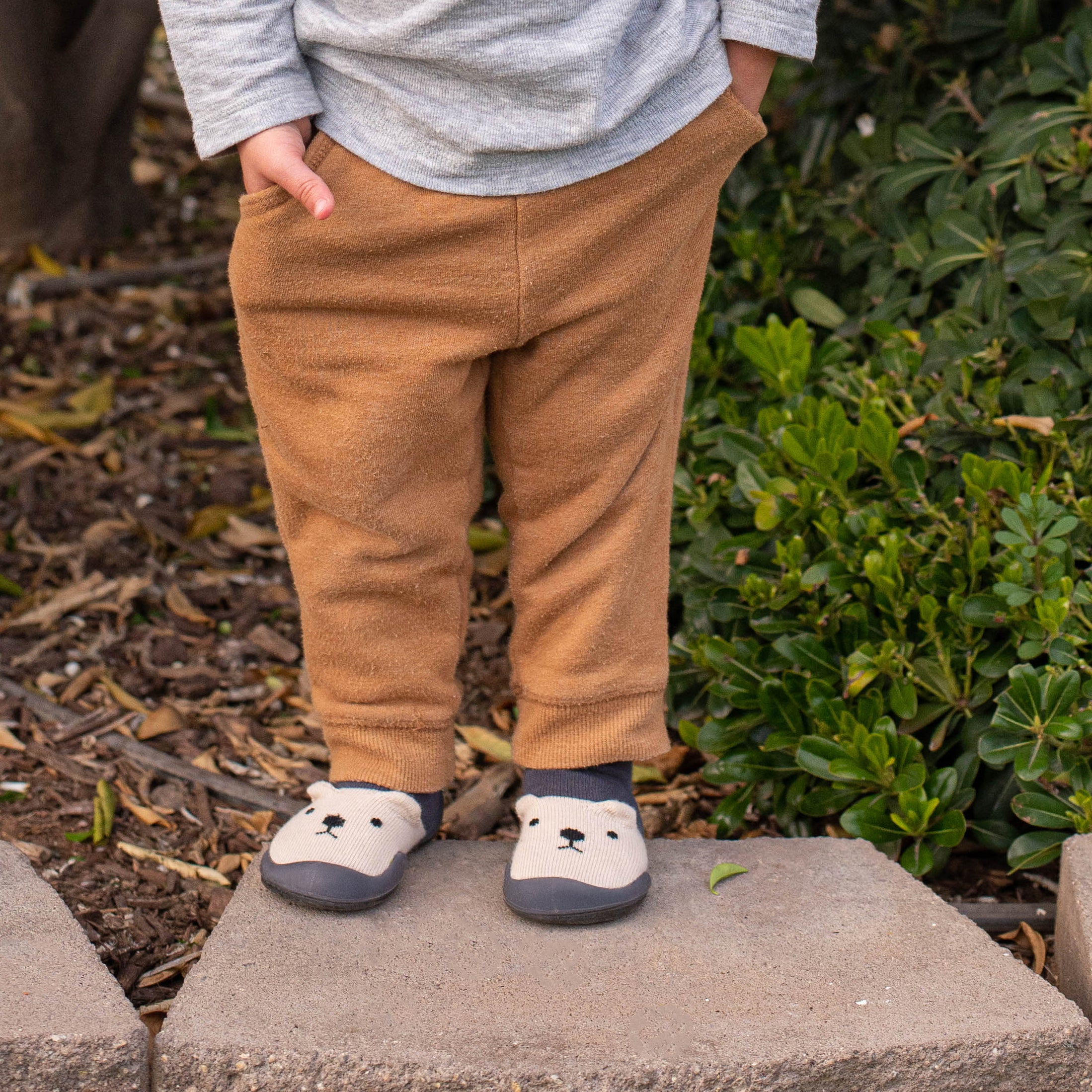 Toddler wearing gray top, brown pants, and bear-themed socks standing on a stone step.
