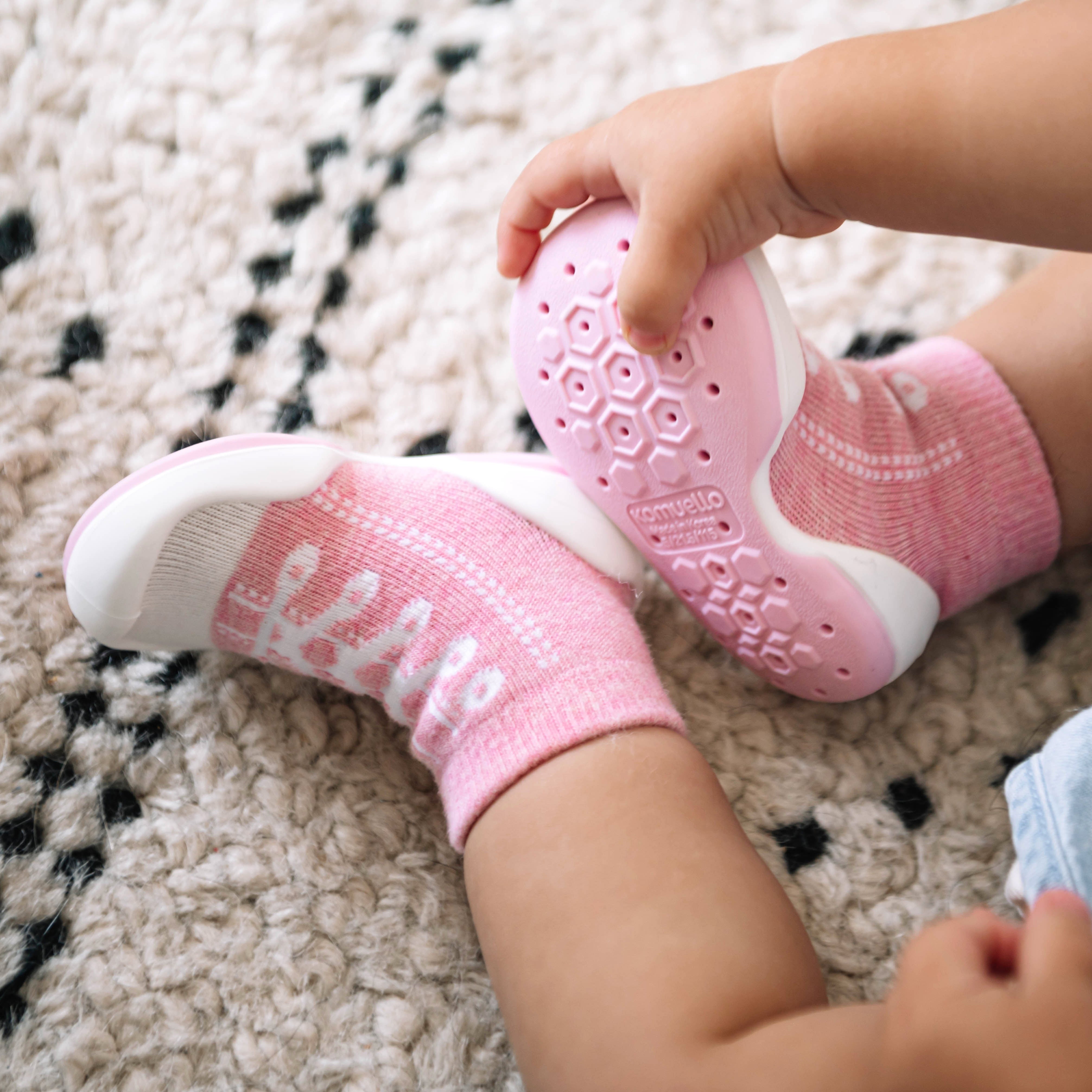 Close-up of a baby's legs and feet in pink Komuello socks shoes with a honeycomb pattern on the sole, resting on a textured rug.