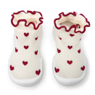 Pair of baby slipper socks, cream colored with red hearts and ruffled trim, white soles.

